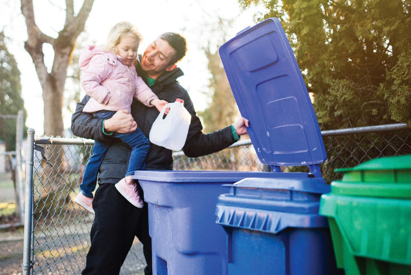 A photo of  a man holding his daughter putting something into the recycling container