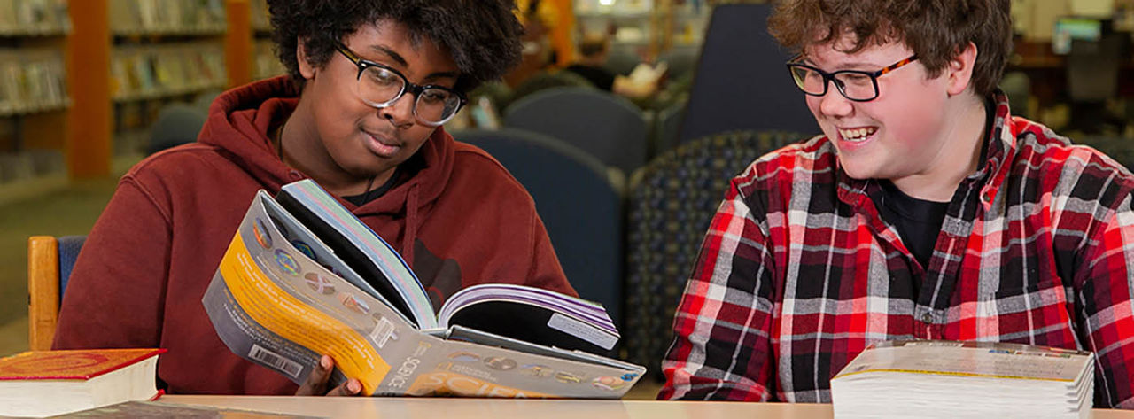 Two teenage boys reading a book in a library study area.