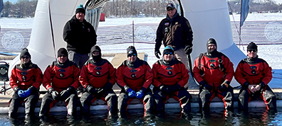 Dive team in red diving gear sitting side by side on a dock in winter.
