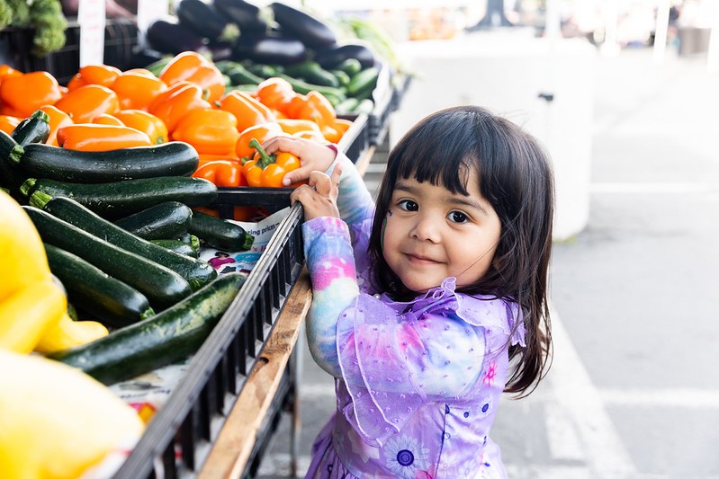 Child at farmers market next to vegetables.