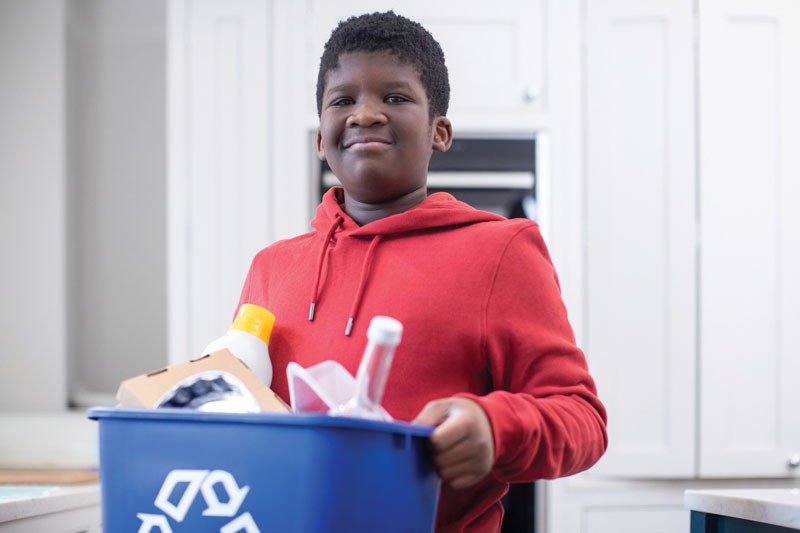Portrait Of Boy Standing In Kitchen At Home Carrying Recycling Bin