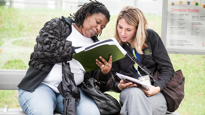 Two people sitting on a bus stop bench, looking at a schedule.