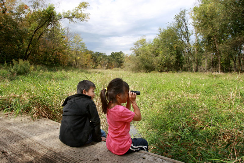 Two children sitting looking out at a grassy area. One child is using binoculars. 