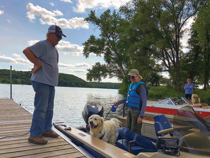 A man standing on a dock next to a boat in the water with a dog and a woman aboard. A second boat is nearby with two passengers aboard.