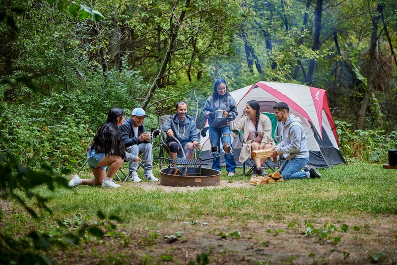A group of people sits around a campfire with a tent in the background.