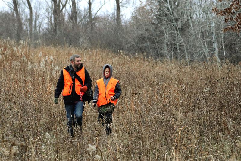 A man and a boy wearing blaze-orange vests walking through a field.