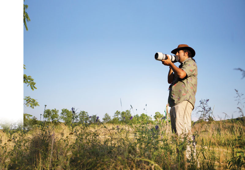 A man standing in a prairie holding a camera with a telephoto lens.