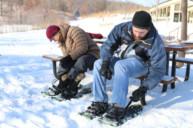 A woman and a man putting on snowshoes while seated at a picnic table.