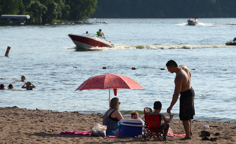 People relaxing on the beach while other people swim and play in the nearby lake with boats in the distance.