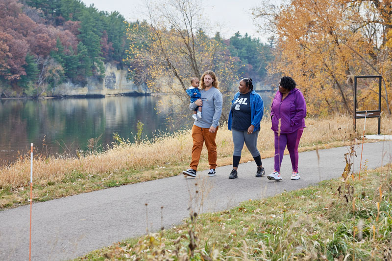 Three adults, one carrying a baby, walking along a paved trail with a lake in the background.