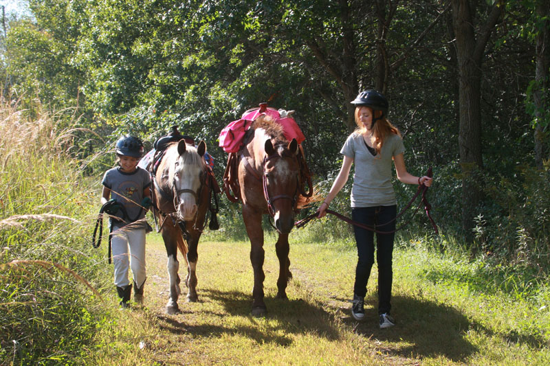 Two teen girls walking horses down a trail. 