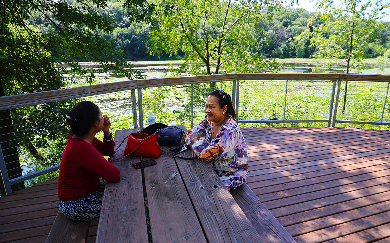A woman smiles at another woman while sitting at a lakeside picnic table.