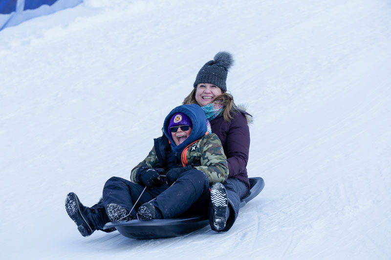 A boy and a woman sliding down a snowy hill on a sled.