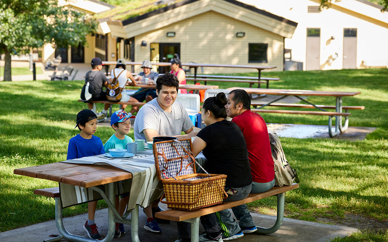 A group of people seated at a picnic table with other picnickers and buildings in the background.
