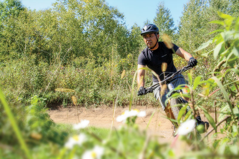 A man riding on a mountain bike trail with plants in the foreground.