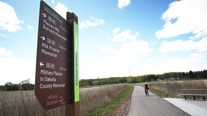 A sign pointing to places of interest on a bike trail with a bicyclist in the background.