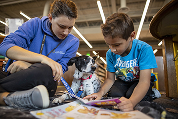 Boy reading a book with a dog next to him. Adult is looking over his shoulder. 